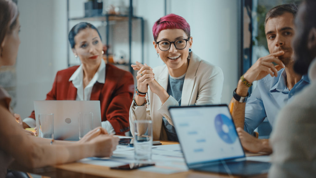 A photo of several people at work having a meeting around a table with their laptops, discussing pronouns.